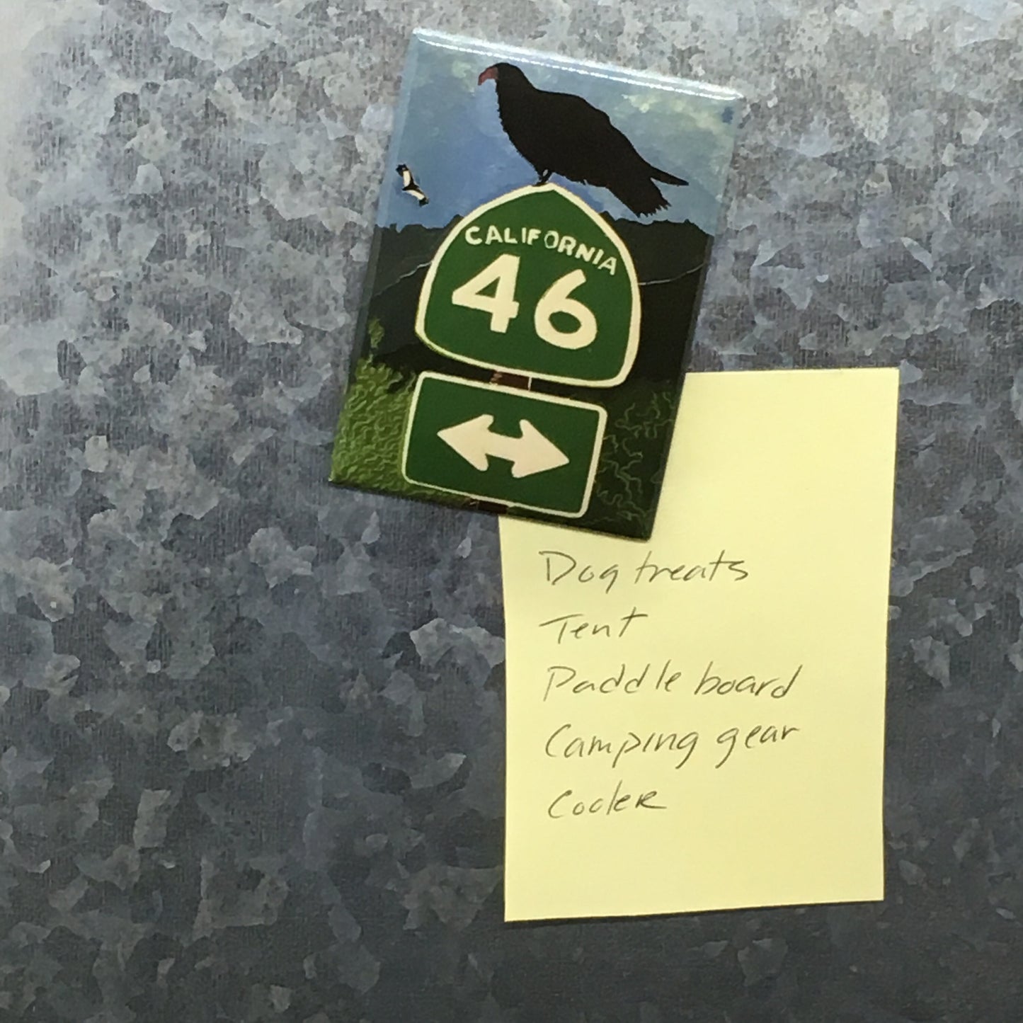 Mylar/metal magnet holding a camping list to a metal surface. Magnet design is a linocut of a turkey vulture perched on a California Highway 46 sign and second vulture flying over hillside in the distance.