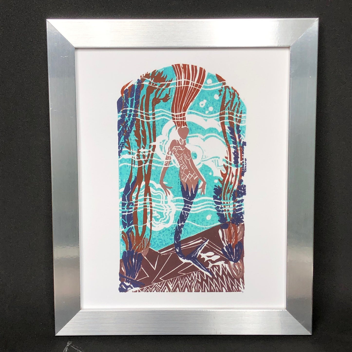 Casino mermaid giclée print of original 5-plate linocut depicting an art deco tile mosaic of the Catalina Casino Mermaid. Bright turquoise back ground with white water movement, stylized mermaid's purple tail surrounded by stylized kelp, coral, and fish. Print is in a silver frame.