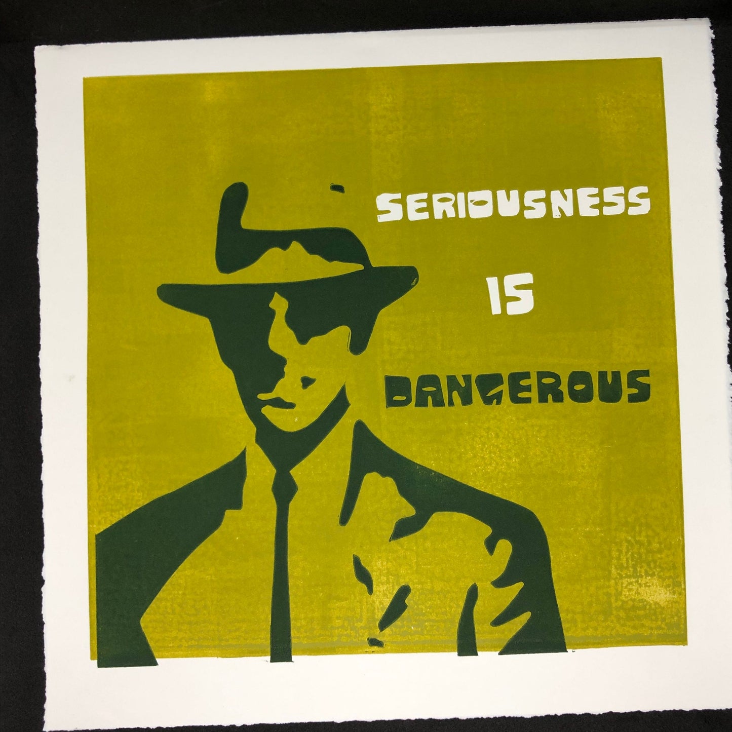 White and forest green text reads: 'Seriousness is Dangerous' to the right of a midcentury modern man dressed up in a suit and tie, donning a fedora hat. The man is forest green on olive green background.