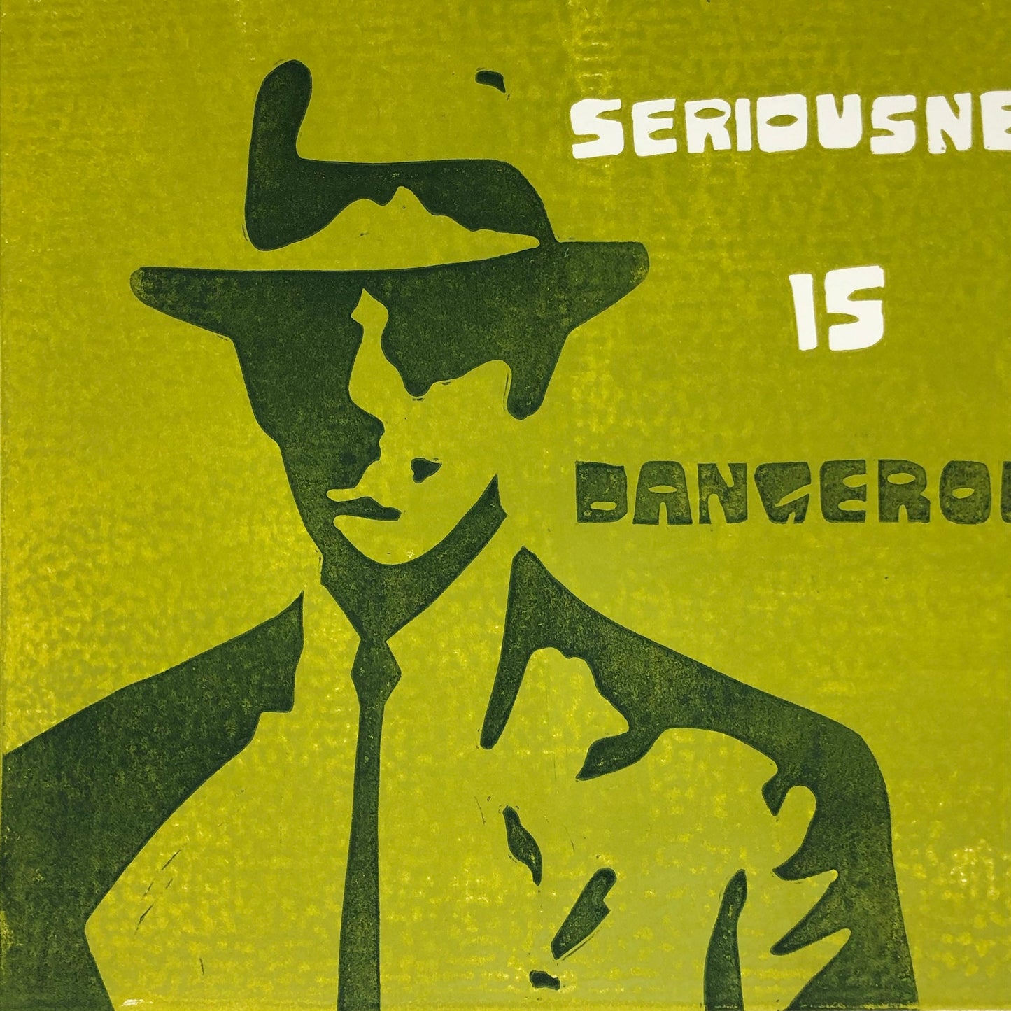 Zoomed in view of print with text partially cut off. White and forest green text reads: 'Seriousness is Dangerous' to the right of a midcentury modern man dressed up in a suit and tie, donning a fedora hat.  The man is forest green on olive green background.